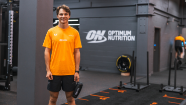 man in orange t-shirt standing in a gym with optimum nutrition wrote on the wall