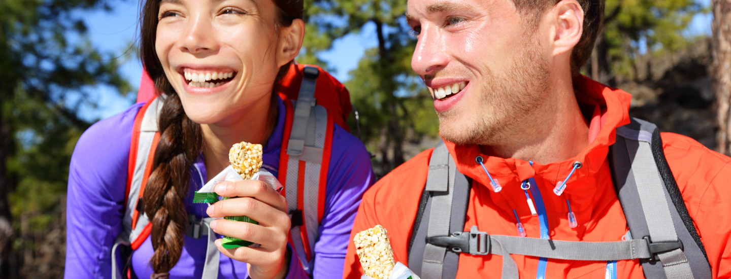 2 people wearing backpacks eating bars