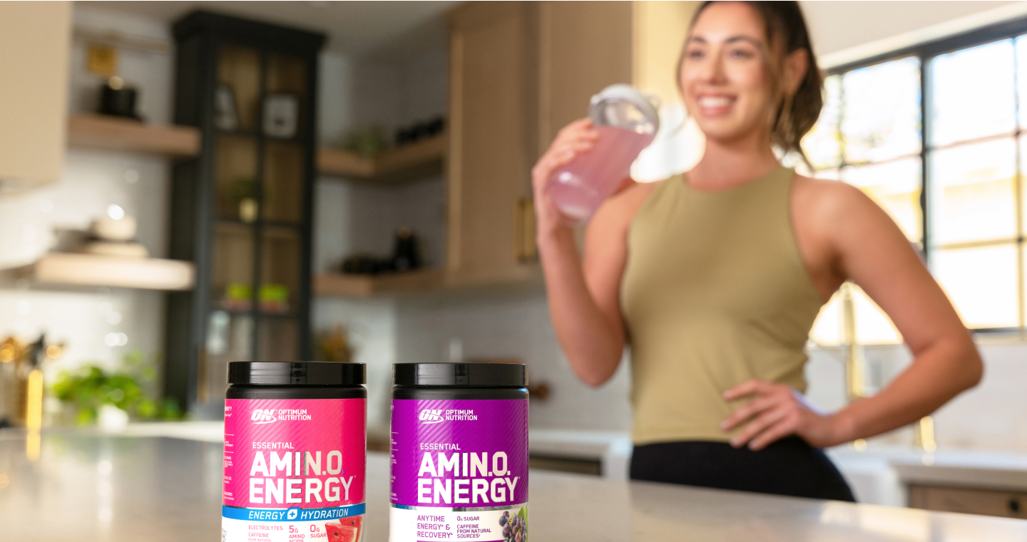 two tubs of optimal nutrition amino energy on table with woman drinking from a bottle behind them