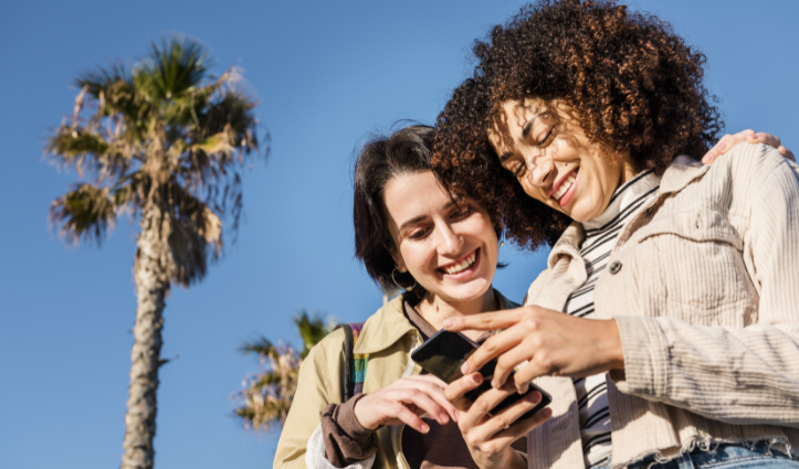Two people standing outdoors under palm trees, looking at a smartphone together.